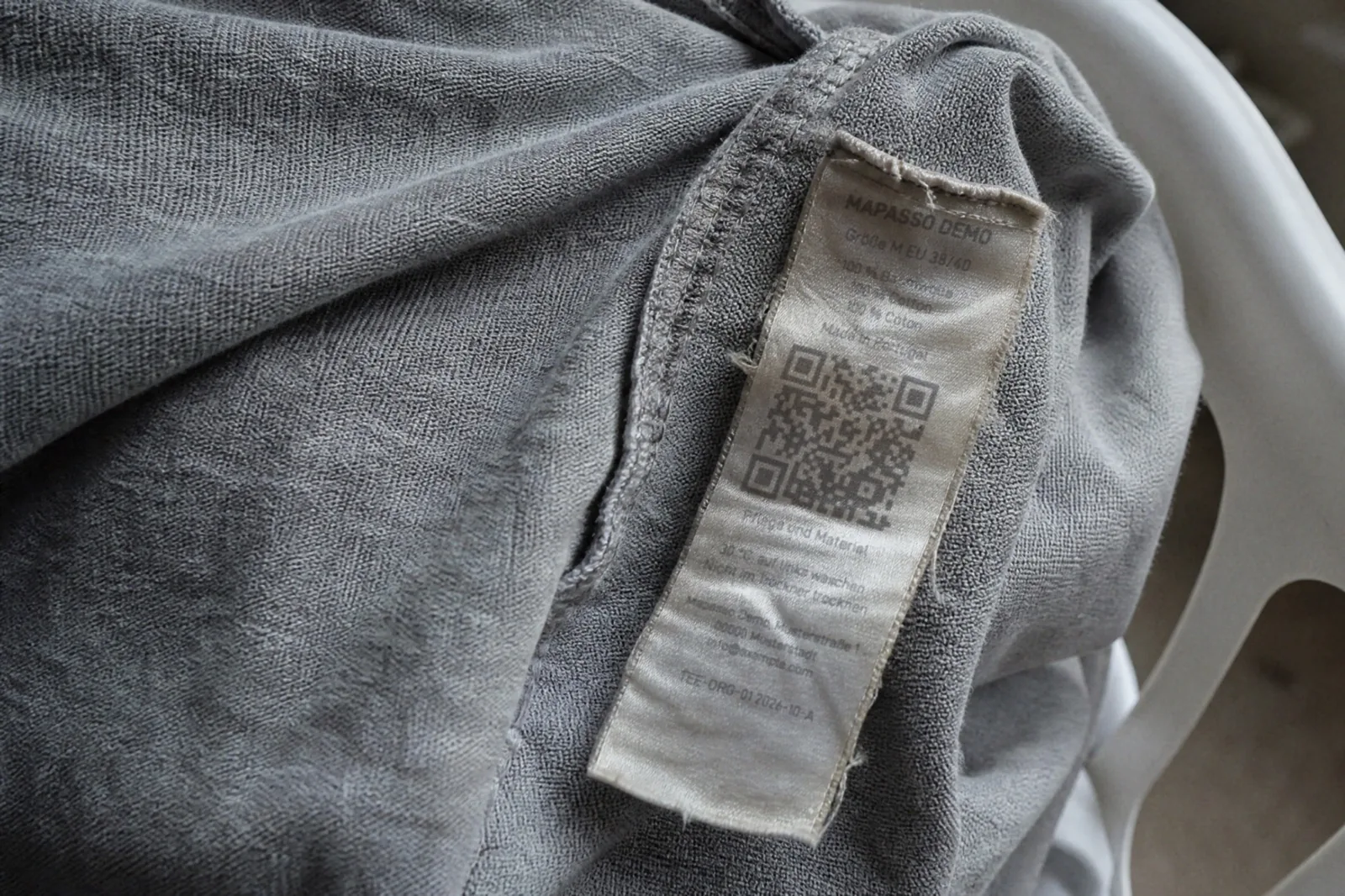 KI-generiertes Bild: Ausgewaschenes, ausgefranstes Pflegeetikett in einem grauen Shirt mit verblasstem Druck und noch lesbarem QR-Code
