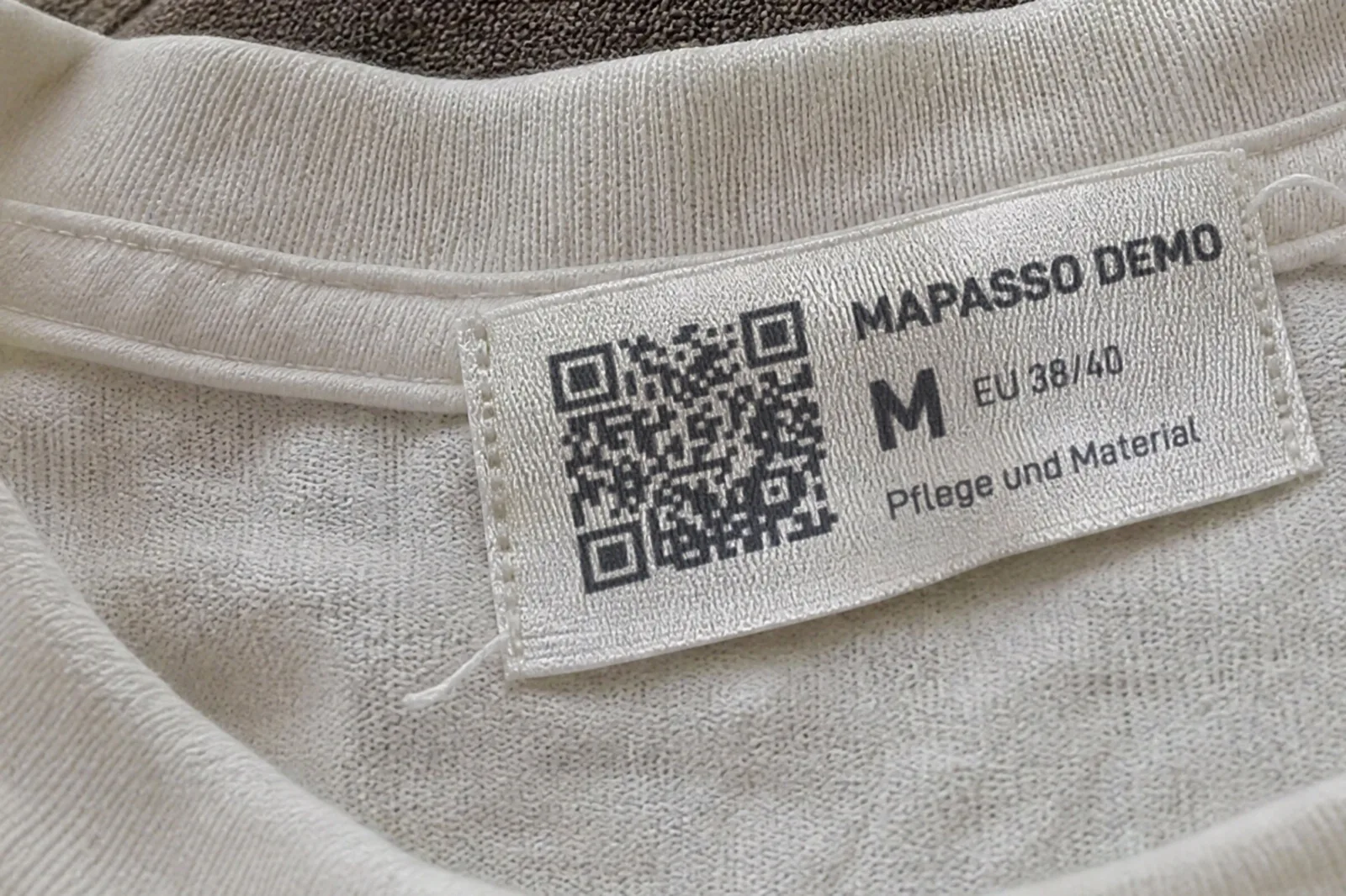 KI-generiertes Bild: Gewebtes Etikett im Nacken eines T-Shirts mit QR-Code, „Mapasso Demo“, Größe M und dem Text „Pflege und Material“