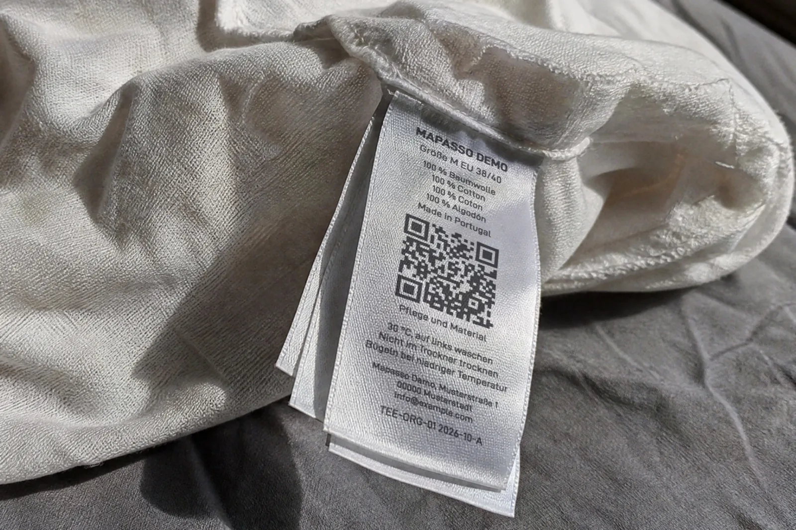KI-generiertes Bild: Pflegeetikett in einem weißen T-Shirt mit QR-Code, „Mapasso Demo“, Größe M, „100 % Baumwolle“ in vier Sprachen, „Made in Portugal“, Pflegehinweisen und der Artikelnummer „TEE-ORG-01“