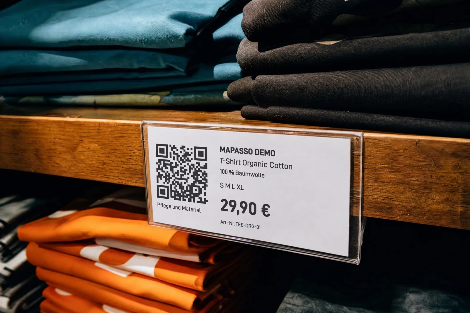 KI-generiertes Bild: Regalschild im Laden mit QR-Code und dem Text „Pflege und Material“, „Mapasso Demo“, „T-Shirt Organic Cotton“, „100 % Baumwolle“, Größen S bis XL und 29,90 €