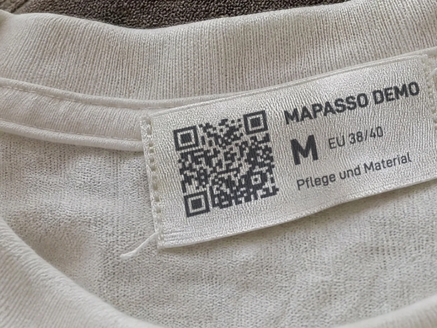 KI-generiertes Bild: Gewebtes Etikett „Mapasso Demo“ mit QR-Code, Größe M und dem Text „Pflege und Material“ im Nacken eines T-Shirts